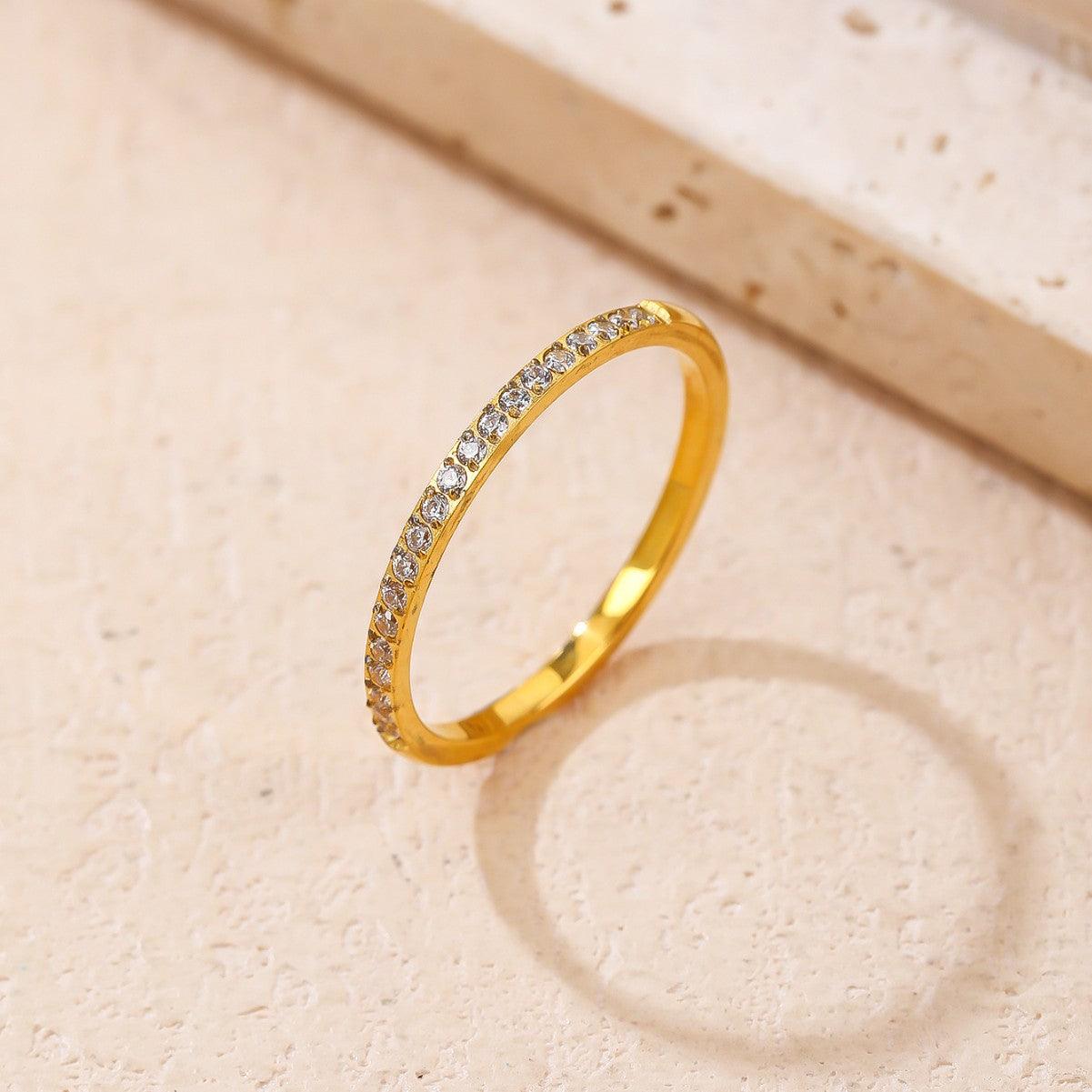 A gold band ring with a row of clear pavé-set stones, photographed on a light beige textured surface with a soft shadow.