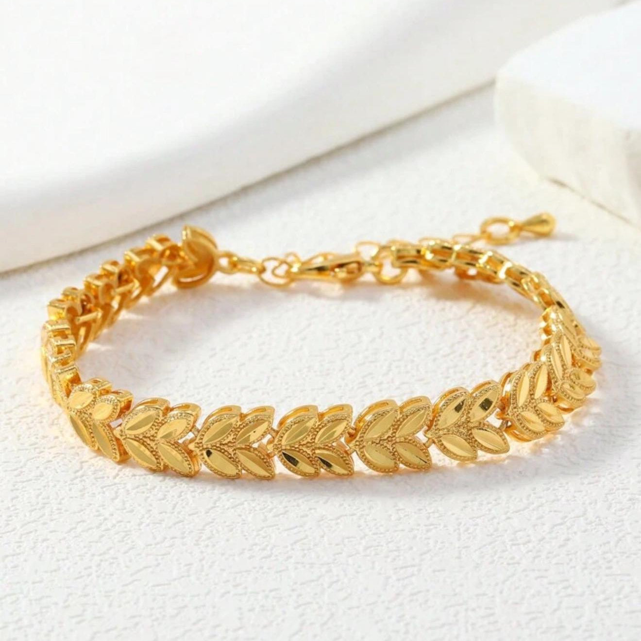 A gold leaf-link bracelet displayed on a textured white surface with a soft white background.
