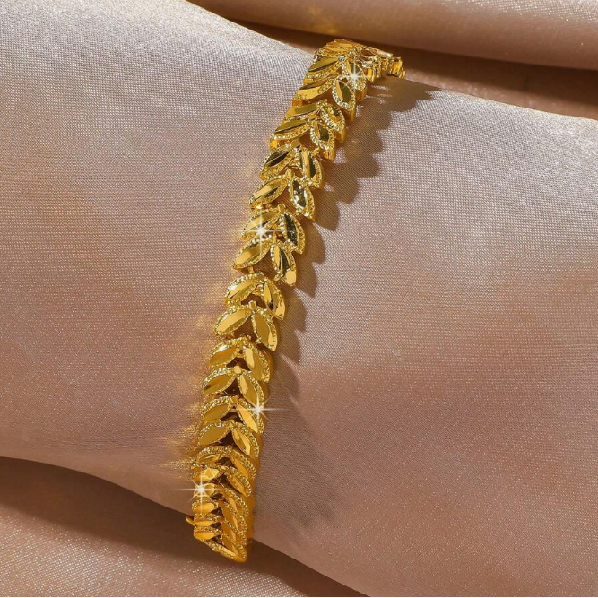 A gold leaf-patterned bangle bracelet displayed on a soft, draped beige fabric background.