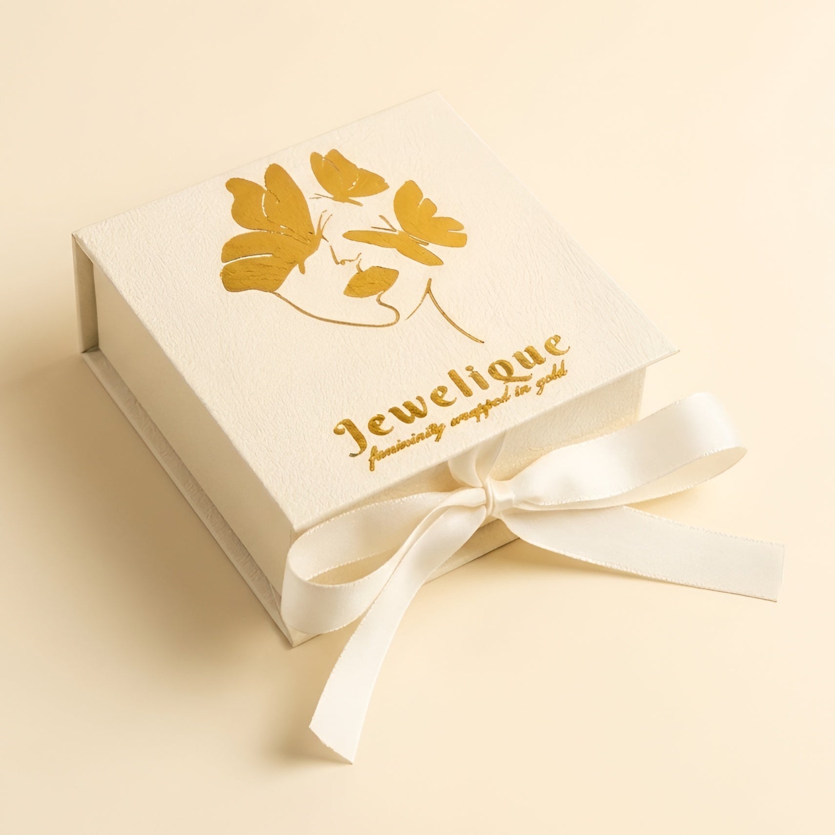 A cream-colored square jewelry gift box with a gold foil butterfly design and the brand name 'Jewelique' printed on the lid, tied with a matching cream ribbon, set against a solid cream background.