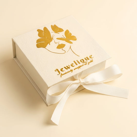 A cream-colored square jewelry gift box with a gold foil butterfly design and the brand name 'Jewelique' printed on the lid, tied with a matching cream ribbon, set against a solid cream background.